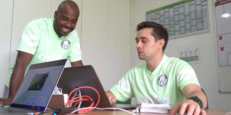 Two men in light green Palmeiras athletic shirts are sitting at a desk in an office, reviewing sports video and data on two open laptops.