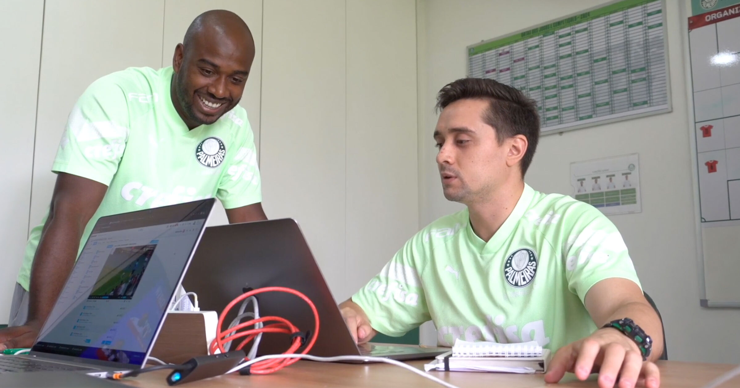 Two men in light green Palmeiras athletic shirts are sitting at a desk in an office, reviewing sports video and data on two open laptops.