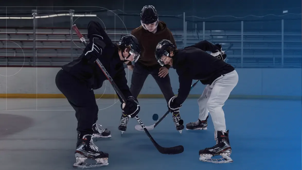 Three hockey players on an ice rink, with two players in black athletic wear facing off over a puck while a third player in a brown hoodie watches from behind.
