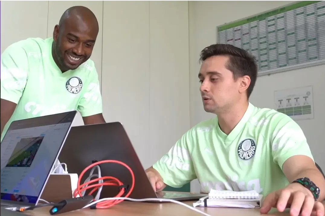 Two men in Palmeiras soccer jerseys look at a laptop screen together, reviewing a soccer game.