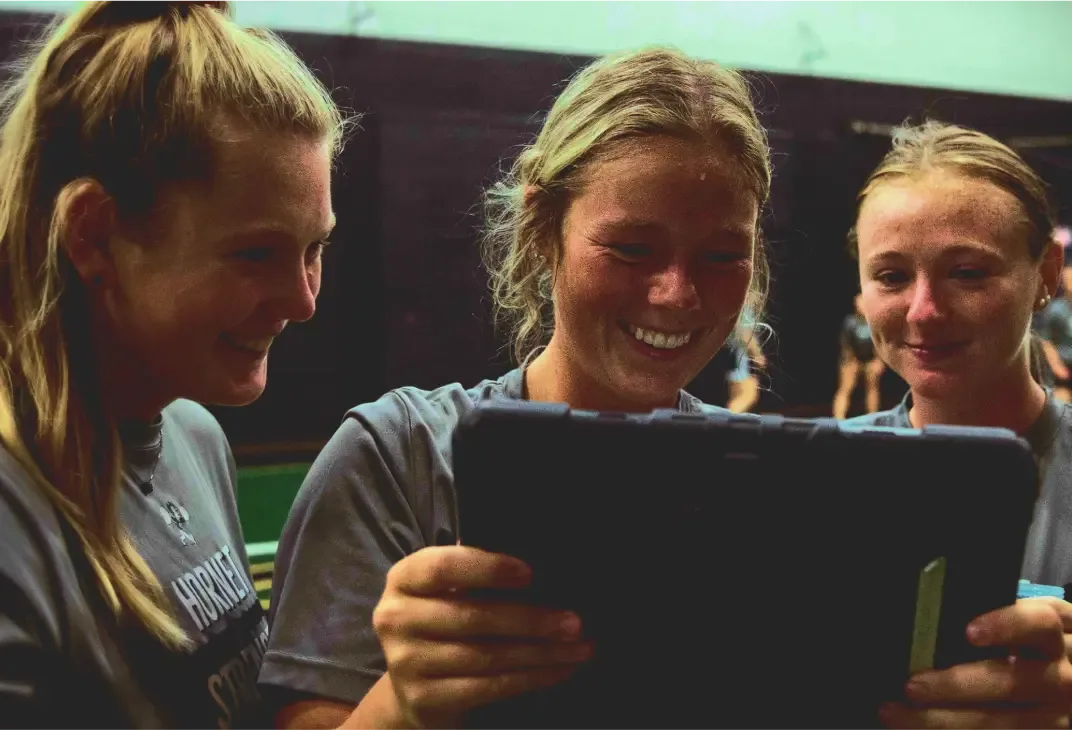 Three female softball players smile while looking at a tablet together.